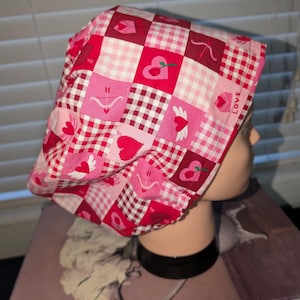 May include: A Valentine's Day themed bouffant scrub cap. The cap features a patchwork design with red, pink, and white squares. Some squares have heart, bow and arrow, and winged heart graphics. The word "LOVE" is printed on the cap.
