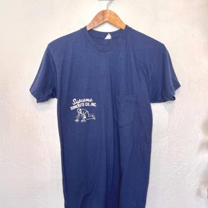 Könnte beinhalten: Marineblaues T-Shirt mit Tasche und dem weißen Text "Supreme Concrete Co, Inc.". Das Shirt hängt an einem Holzbügel vor einer weißen Wand.