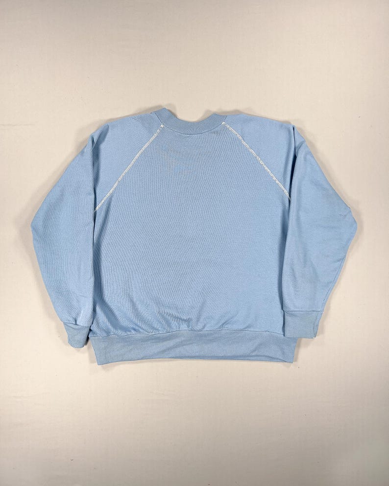 May include: A pale blue sweatshirt with a crew neck and long sleeves. White stitching details the shoulder seams. The cuffs and waistband are a slightly darker shade of blue.