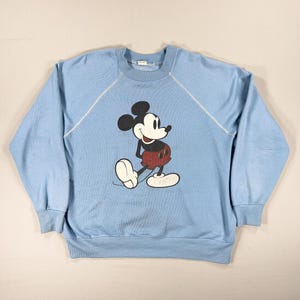 May include: Pale blue sweatshirt with a Mickey Mouse design. The cartoon mouse is in black, red, and white. The long-sleeved top has a round neck and white stitching. A vintage style garment.