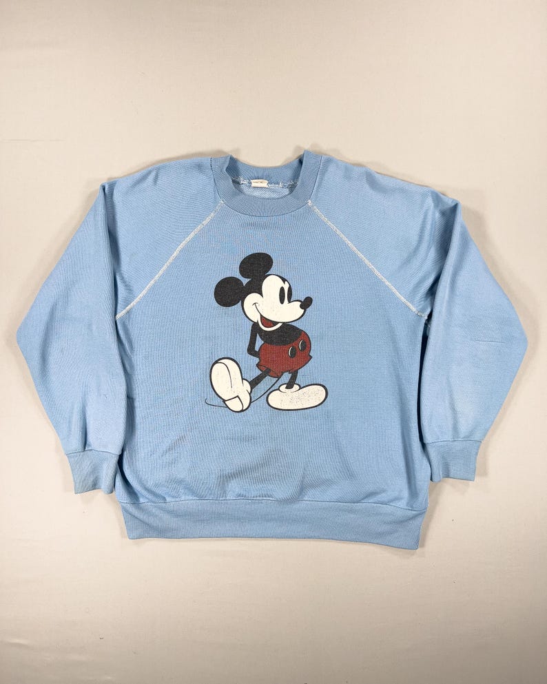 May include: Pale blue sweatshirt with a Mickey Mouse design. The cartoon mouse is in black, red, and white. The long-sleeved top has a round neck and white stitching. A vintage style garment.