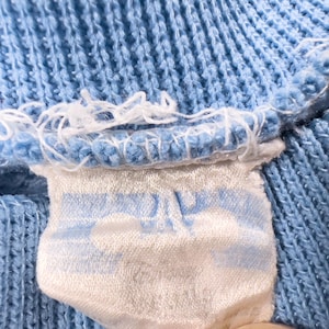 May include: Close-up of a pale blue knitted fabric with a white label attached. The label features a light blue design and is sewn into the fabric. The fabric has a textured appearance.