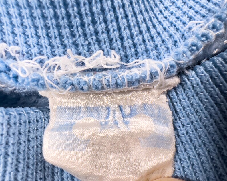 May include: Close-up of a pale blue knitted fabric with a white label attached. The label features a light blue design and is sewn into the fabric. The fabric has a textured appearance.