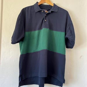 Pode incluir: Uma camisa polo azul marinho e verde com blocos de cores e mangas curtas. A camisa está pendurada em um cabide de madeira. A camisa tem gola e carcela de três botões.