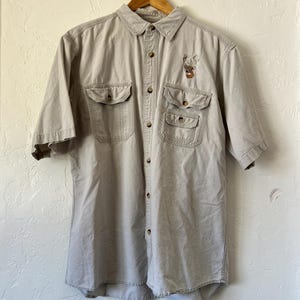 Puede incluir: Una camisa beige de manga corta con dos bolsillos en el pecho y un diseño bordado en el bolsillo izquierdo. La camisa está colgada en una percha de madera contra una pared de color claro.
