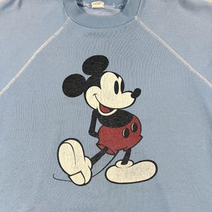 May include: Pale blue sweatshirt with a distressed Mickey Mouse graphic. The cartoon mouse is in black, red, and white, smiling. The sweatshirt has a crew neck and raglan sleeves. A vintage style.