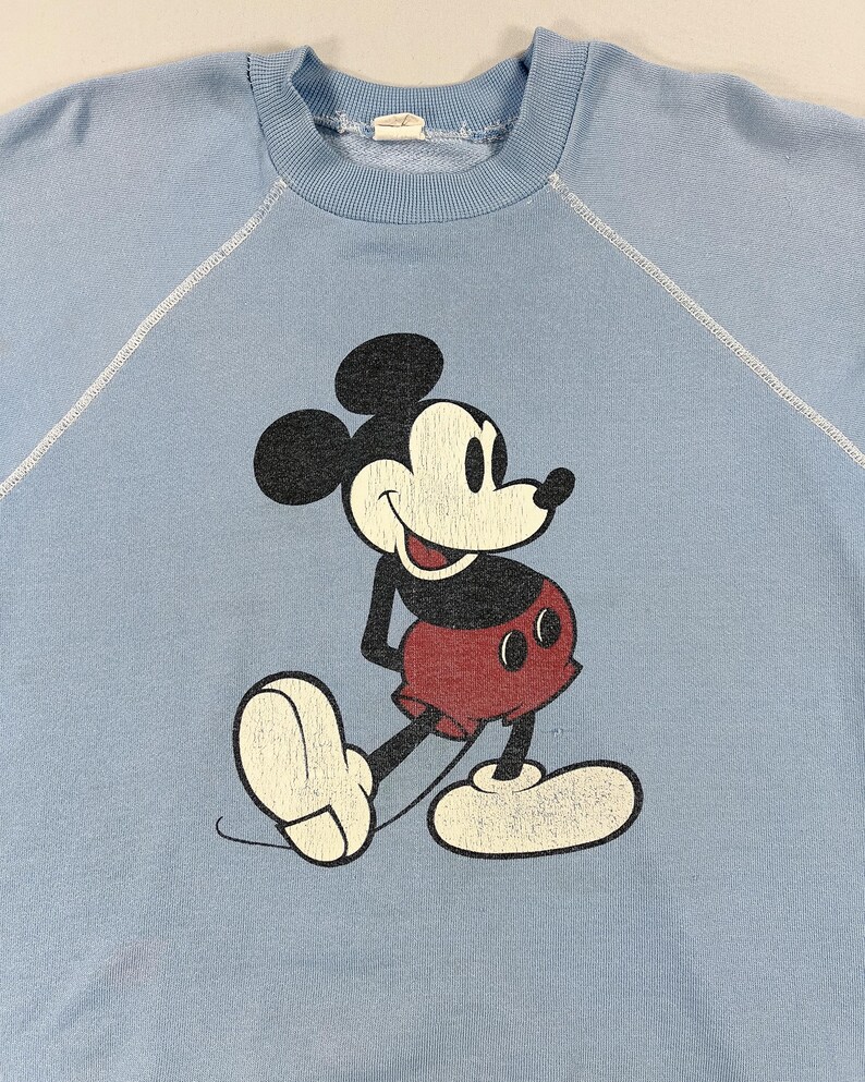 May include: Pale blue sweatshirt with a distressed Mickey Mouse graphic. The cartoon mouse is in black, red, and white, smiling. The sweatshirt has a crew neck and raglan sleeves. A vintage style.