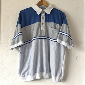 Peut inclure: Un polo vintage avec un design en blocs de couleurs. Le haut présente une section supérieure bleue, une section centrale grise et une section inférieure rayée. La chemise a un col et des poignets blancs et est suspendue à un cintre en bois.
