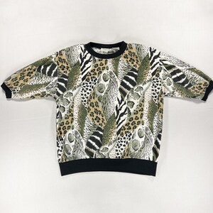 Peut inclure: Un sweat-shirt à manches courtes avec un imprimé animalier audacieux dans les tons de beige, noir et vert olive. L'encolure ras du cou et les poignets des manches sont bordés de noir. Le tissu présente un mélange de motifs léopard, zèbre et autres animaux.