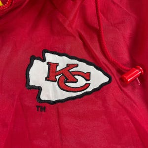 May include: A red nylon jacket with a drawstring hood and a zipper closure. The jacket features the Kansas City Chiefs logo, a white arrowhead with a black outline, and the letters "KC" in red.