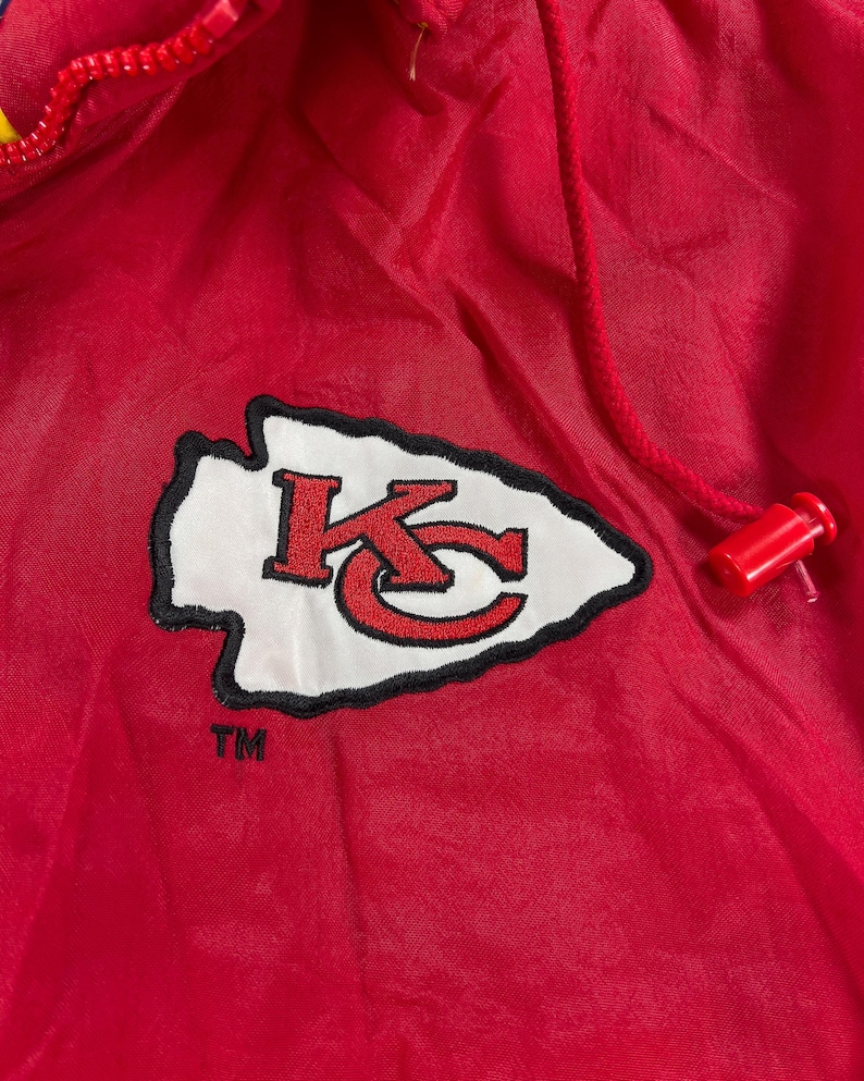 May include: A red nylon jacket with a drawstring hood and a zipper closure. The jacket features the Kansas City Chiefs logo, a white arrowhead with a black outline, and the letters "KC" in red.