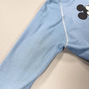 May include: A pale blue sweatshirt featuring a black and white graphic on the upper right. The long sleeve has white stitching along the seams. The fabric appears to be a soft, knit material.