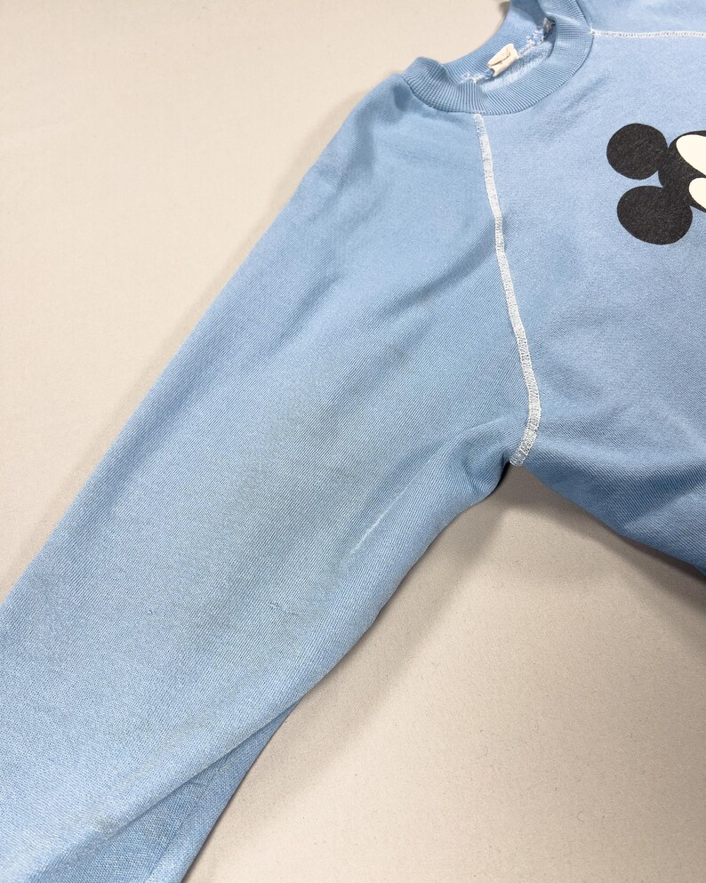 May include: A pale blue sweatshirt featuring a black and white graphic on the upper right. The long sleeve has white stitching along the seams. The fabric appears to be a soft, knit material.