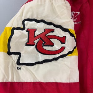 May include: Red and white athletic jacket with a large embroidered logo. The logo features a red "KC" design with a black outline on a white background, bordered by yellow and black stripes. The jacket has the word "ATHLETIC" printed on it.