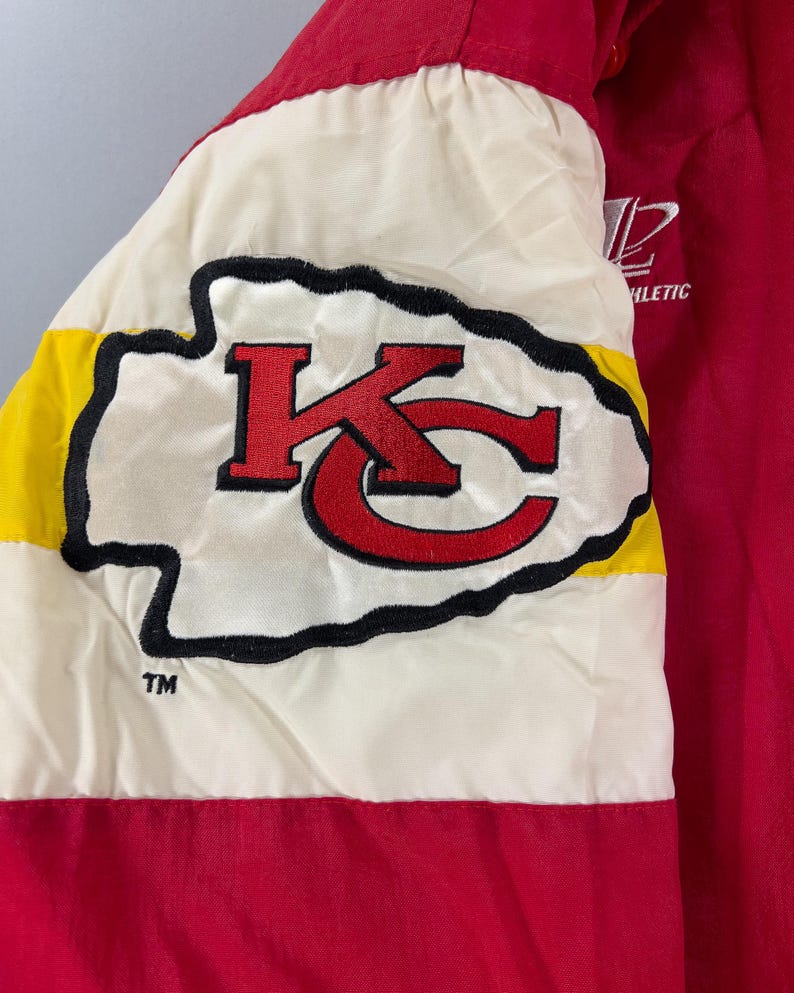 May include: Red and white athletic jacket with a large embroidered logo. The logo features a red "KC" design with a black outline on a white background, bordered by yellow and black stripes. The jacket has the word "ATHLETIC" printed on it.