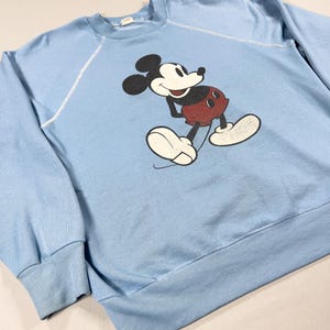 May include: A pale blue sweatshirt with a Mickey Mouse graphic. The cartoon character is in black, white, and red. The sweatshirt has a crew neck, long sleeves, and ribbed cuffs and hem. The stitching is white.