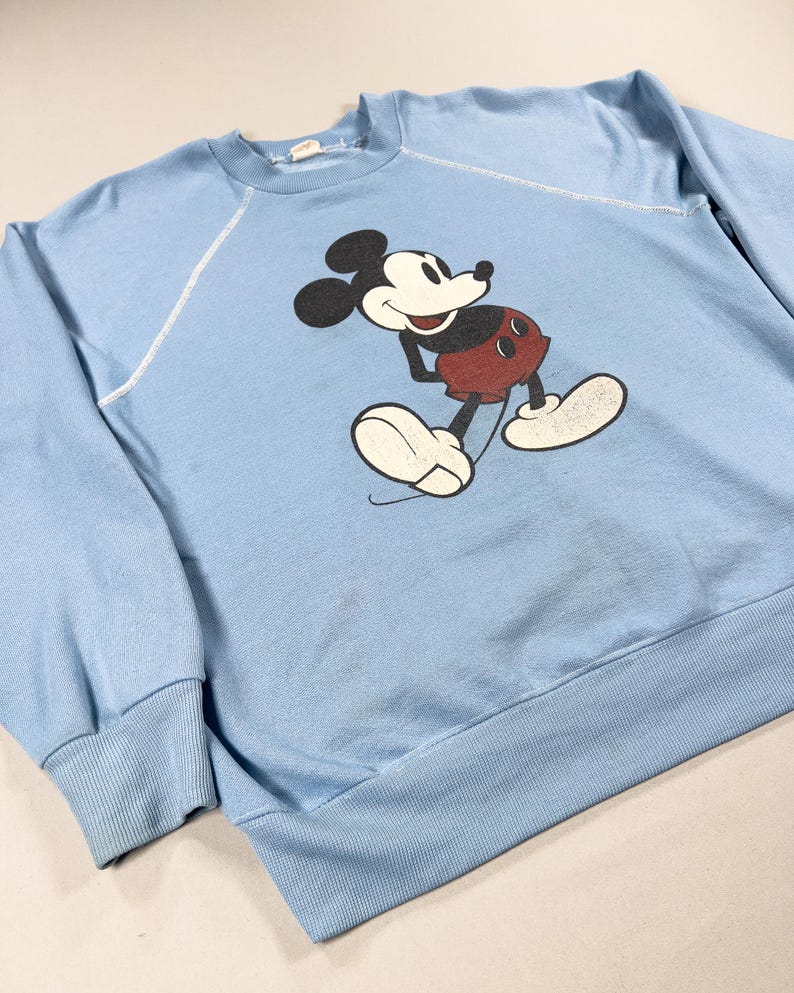 May include: A pale blue sweatshirt with a Mickey Mouse graphic. The cartoon character is in black, white, and red. The sweatshirt has a crew neck, long sleeves, and ribbed cuffs and hem. The stitching is white.