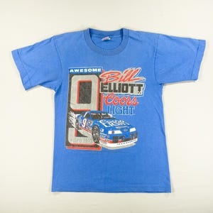 以下が含まれることがあります： 水色のTシャツ。レーシングカーのグラフィック、ナンバー9、そして「Bill Elliott Coors Light」の文字がプリントされています。「AWESOME」の文字がナンバー9の上にプリントされています。