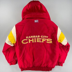 May include: Red hooded jacket with the text "Kansas City Chiefs" in gold lettering. The jacket has yellow and white accents on the sleeves and a "Pro Line" logo. The jacket is a vintage style.
