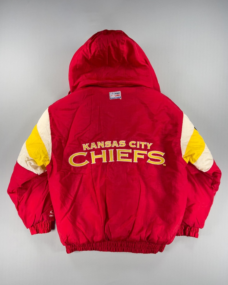 May include: Red hooded jacket with the text "Kansas City Chiefs" in gold lettering. The jacket has yellow and white accents on the sleeves and a "Pro Line" logo. The jacket is a vintage style.
