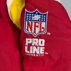 May include: Close-up of a red, yellow, and white jacket. The jacket features an NFL logo patch with the text "NFL" and "PRO LINE AUTHENTIC" in white embroidery. The jacket is made of a shiny, possibly nylon, material.
