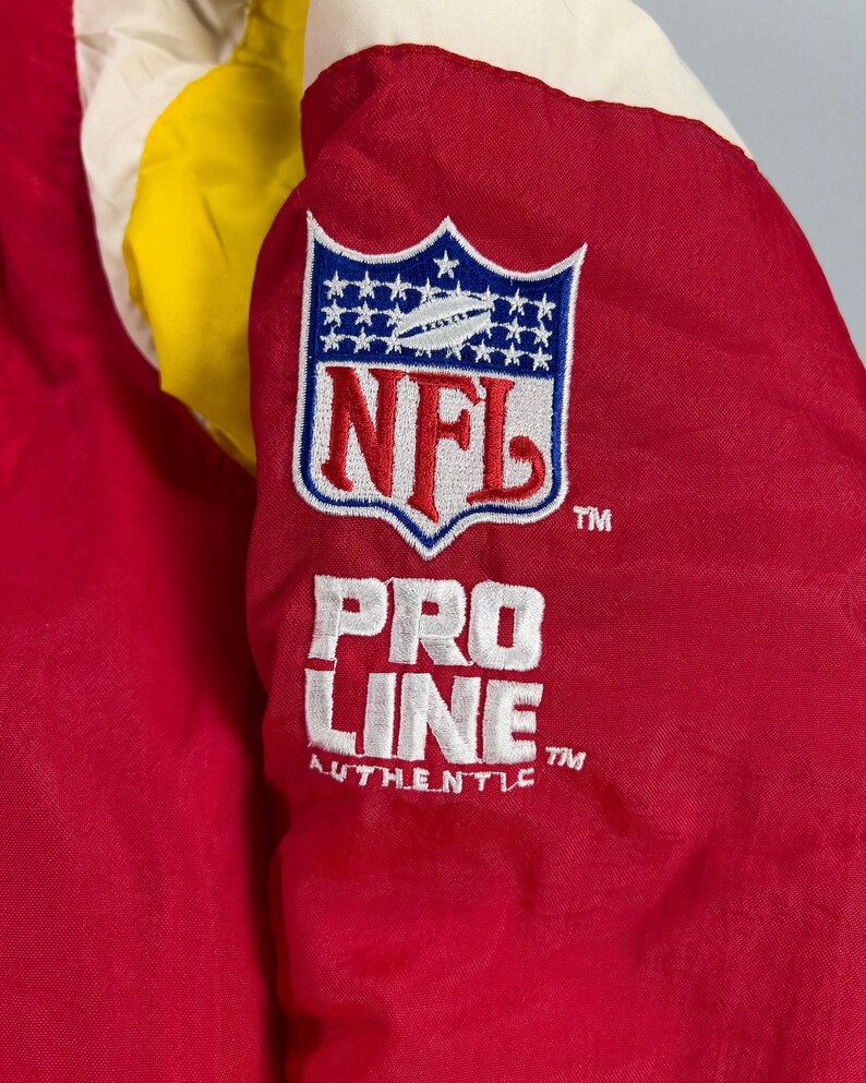 May include: Close-up of a red, yellow, and white jacket. The jacket features an NFL logo patch with the text "NFL" and "PRO LINE AUTHENTIC" in white embroidery. The jacket is made of a shiny, possibly nylon, material.