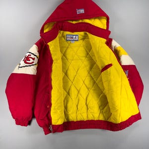 May include: Red and yellow hooded jacket with a quilted yellow interior. The jacket features a red hood, sleeves, and body with white and red accents. The left sleeve displays a team logo. The jacket has a zipper closure and a red ribbed hem.
