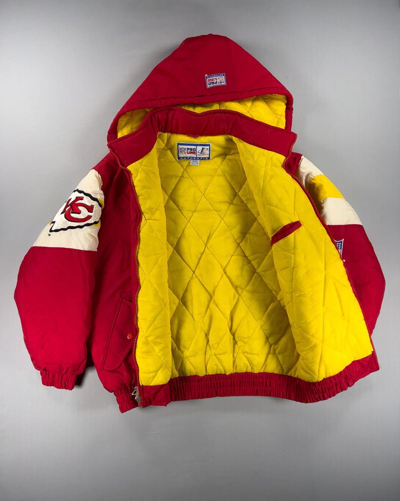 May include: Red and yellow hooded jacket with a quilted yellow interior. The jacket features a red hood, sleeves, and body with white and red accents. The left sleeve displays a team logo. The jacket has a zipper closure and a red ribbed hem.