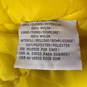 May include: A close-up of a white tag with black text, set against a bright yellow quilted fabric. The tag lists the materials: 100% nylon for the shell and lining, and 100% polyester for the interfill. The tag also includes care instructions.