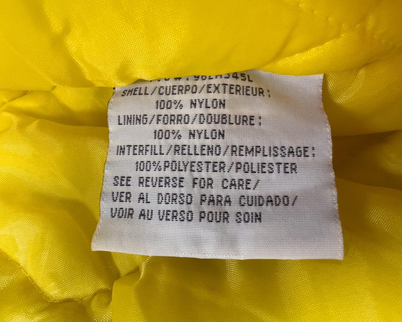 May include: A close-up of a white tag with black text, set against a bright yellow quilted fabric. The tag lists the materials: 100% nylon for the shell and lining, and 100% polyester for the interfill. The tag also includes care instructions.