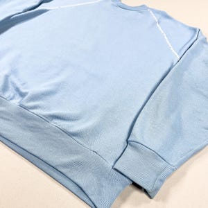 May include: A pale blue sweatshirt featuring a ribbed hem and cuffs. White stitching accents the raglan sleeves. The fabric looks like a soft knit, ideal for casual wear. The design is simple and classic, suitable for everyday use.