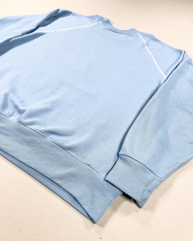May include: A pale blue sweatshirt featuring a ribbed hem and cuffs. White stitching accents the raglan sleeves. The fabric looks like a soft knit, ideal for casual wear. The design is simple and classic, suitable for everyday use.