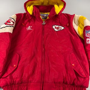 May include: A red hooded jacket with yellow lining and white and yellow accents on the sleeves. The jacket features the Kansas City Chiefs logo and NFL Pro Line branding. It has a full zipper and two front pockets.