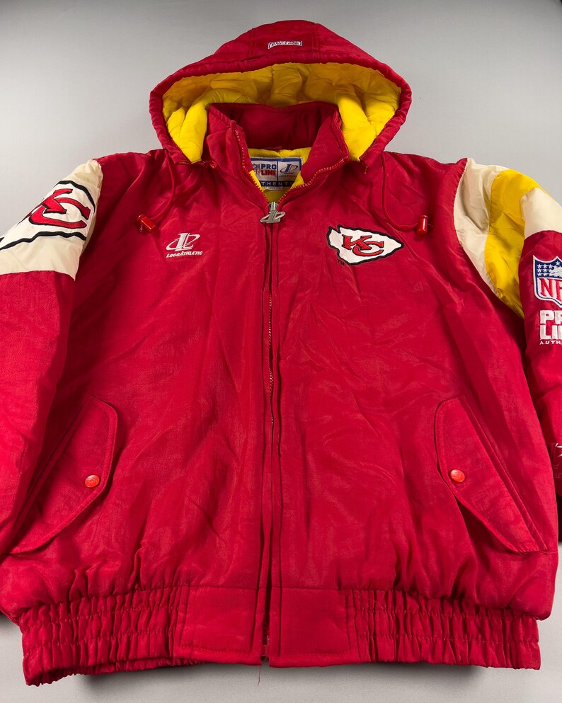 May include: A red hooded jacket with yellow lining and white and yellow accents on the sleeves. The jacket features the Kansas City Chiefs logo and NFL Pro Line branding. It has a full zipper and two front pockets.