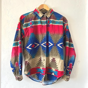 Puede incluir: Una camisa de manga larga con un colorido estampado geométrico. La camisa presenta una mezcla de colores rojo, azul, marrón y beige. El diseño incluye formas de diamantes y zigzags. La camisa está colgada en una percha de madera.