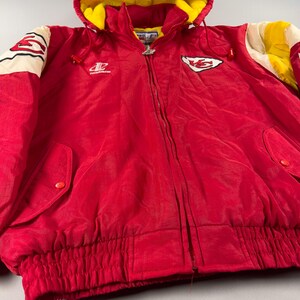 May include: Red hooded jacket with yellow lining and white and yellow accents. Features a full zipper, elastic waist, and the Kansas City Chiefs logo on the chest. The sleeves have white and yellow stripes. A vintage sports jacket.