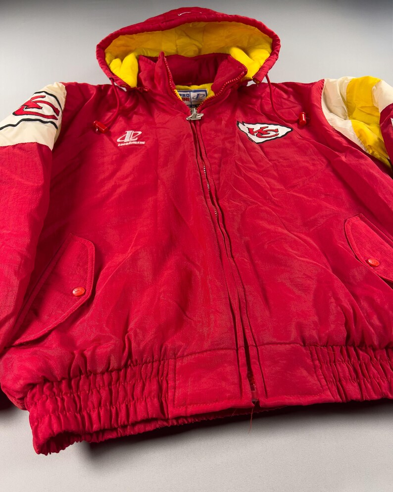 May include: Red hooded jacket with yellow lining and white and yellow accents. Features a full zipper, elastic waist, and the Kansas City Chiefs logo on the chest. The sleeves have white and yellow stripes. A vintage sports jacket.