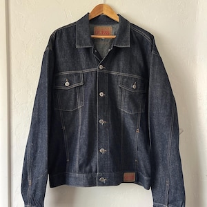 May include: Dark wash denim jacket with the "GUESS" brand label. The jacket features a classic design with button closures, two chest pockets, and two side pockets. The jacket is hanging on a wooden hanger.
