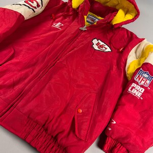 May include: A red hooded jacket with a yellow interior lining. The jacket features the Kansas City Chiefs logo on the chest and sleeve, along with the NFL Pro Line logo. The jacket has a zipper closure and two front pockets.