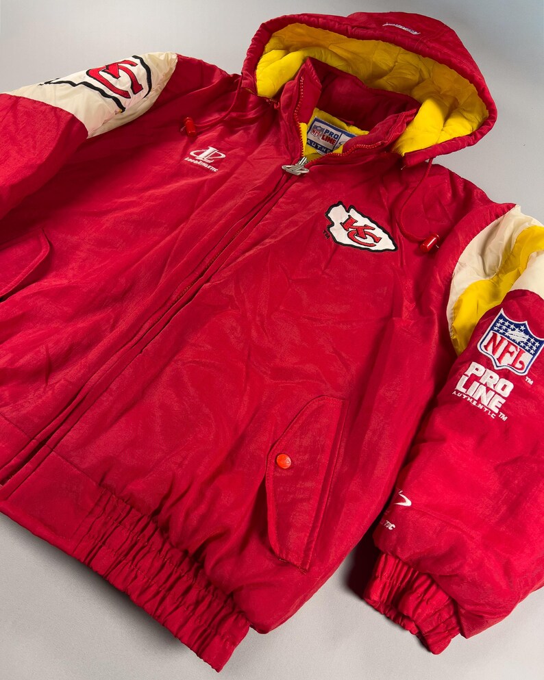 May include: A red hooded jacket with a yellow interior lining. The jacket features the Kansas City Chiefs logo on the chest and sleeve, along with the NFL Pro Line logo. The jacket has a zipper closure and two front pockets.