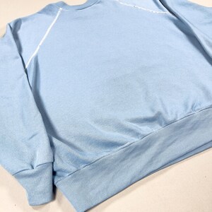 May include: A pale blue sweatshirt with a crew neck and long sleeves. White stitching details the shoulder seams. It features a ribbed hem and cuffs. The fabric looks soft and comfortable.