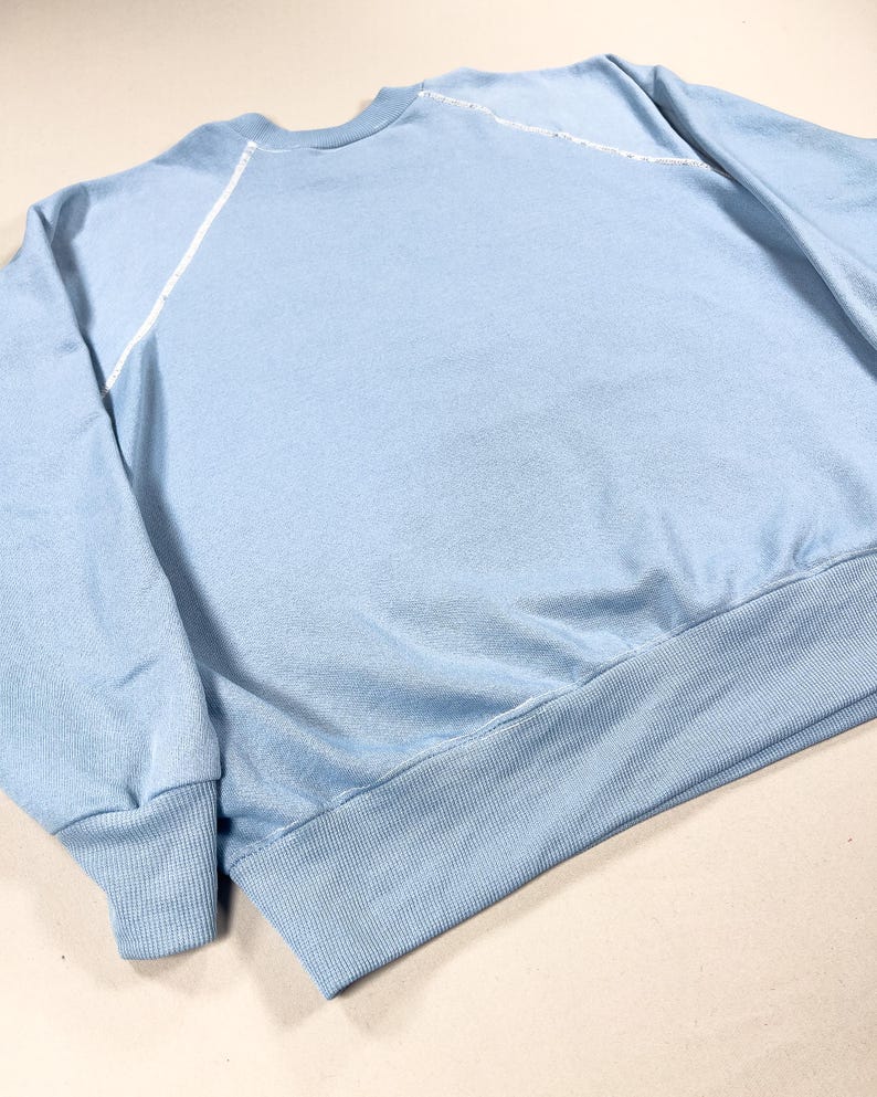 May include: A pale blue sweatshirt with a crew neck and long sleeves. White stitching details the shoulder seams. It features a ribbed hem and cuffs. The fabric looks soft and comfortable.