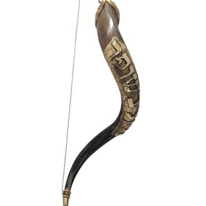 May include: A curved shofar horn featuring a polished dark brown and translucent finish, adorned with ornate gold-toned metal plating that displays Hebrew lettering along its length. A thin, light-colored string is attached to the tip and base.