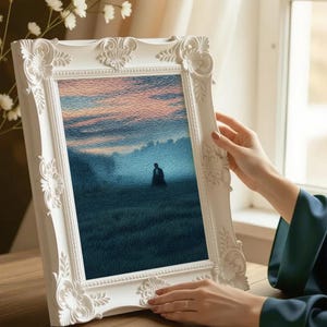 May include: A white, ornate picture frame holding a landscape painting of a person in a field. The painting features a blue and pink sky and a figure in the distance. The frame is held by a person with a dark green sleeve.