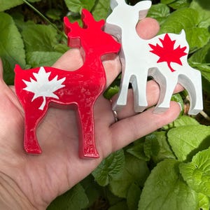 Peut inclure: Deux figurines de cerf, une rouge et une blanche, chacune avec un motif de feuille d'érable. Le cerf rouge a une feuille d'érable blanche, tandis que le cerf blanc a une feuille d'érable rouge. Les figurines sont posées sur un fond de feuillage vert.