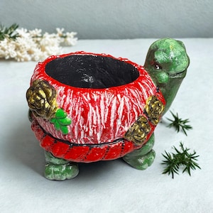 Könnte beinhalten: Keramik-Schildkröten-Pflanzgefäß mit rotem und weißem Muster auf dem Panzer und grünen Kopf und Beinen. Das Pflanzgefäß hat einen schwarzen Innenraum und ist mit goldfarbenen floralen Akzenten verziert. Ein dekorativer Artikel für die Inneneinrichtung.