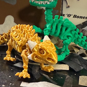 Puede incluir: Dos modelos de dinosaurios impresos en 3D. Uno es dorado y blanco, parecido a un lagarto, y el otro es verde, esquelético y erguido. El dinosaurio verde tiene un cráneo y una caja torácica detallados. El dinosaurio dorado está en posición de gateo.