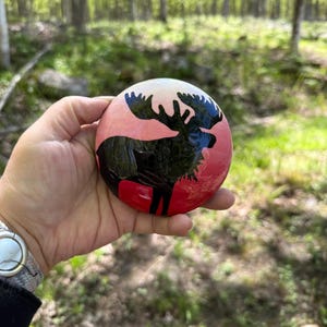 May include: A hand-painted, round stone featuring a black moose silhouette against a gradient background of pink and white. The moose has large antlers and is set against a red base. The stone is held outdoors with a forest backdrop.