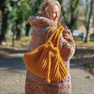 May include: A mustard yellow, hand-knitted tote bag with thick, textured yarn. The bag is held by a person wearing a hooded, oversized sweater in a multicolored knit pattern of orange, grey, and red. The background is a blurred outdoor scene.