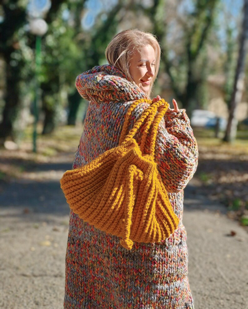 May include: A mustard yellow, hand-knitted tote bag with thick, textured yarn. The bag is held by a person wearing a hooded, oversized sweater in a multicolored knit pattern of orange, grey, and red. The background is a blurred outdoor scene.